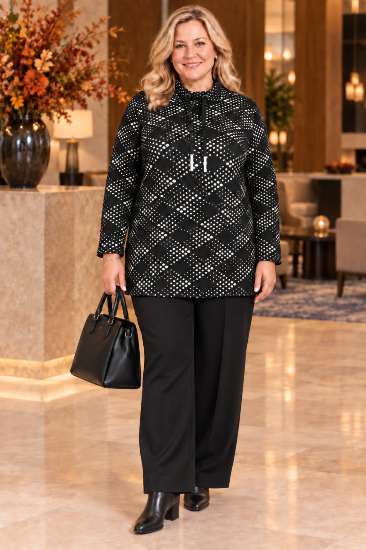A woman models a black long-sleeve top with a white argyle pattern, paired with black wide-leg pants and holding a black handbag in a luxurious indoor lobby setting.