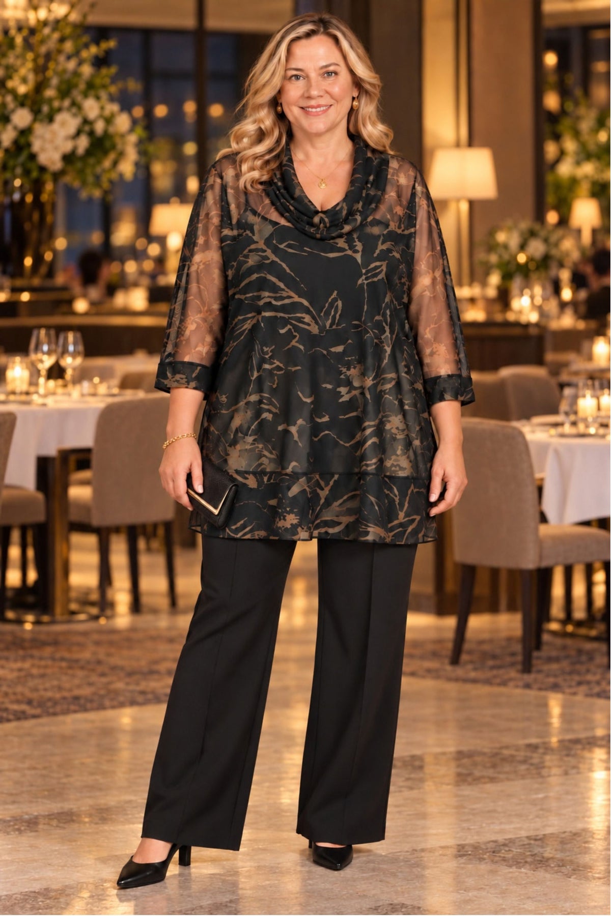A woman stands in a restaurant wearing a sheer black tunic top with a metallic gold abstract pattern, paired with black wide-leg trousers and black heels.
