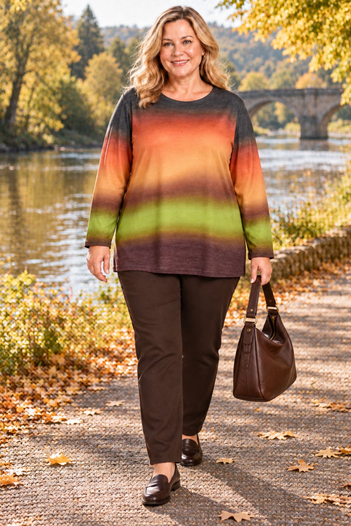 A woman walking outdoors on a path by a river, wearing a long-sleeve crewneck top with horizontal stripes in dark gray, orange, yellow, and olive green, paired with dark brown pants and carrying a brown handbag.