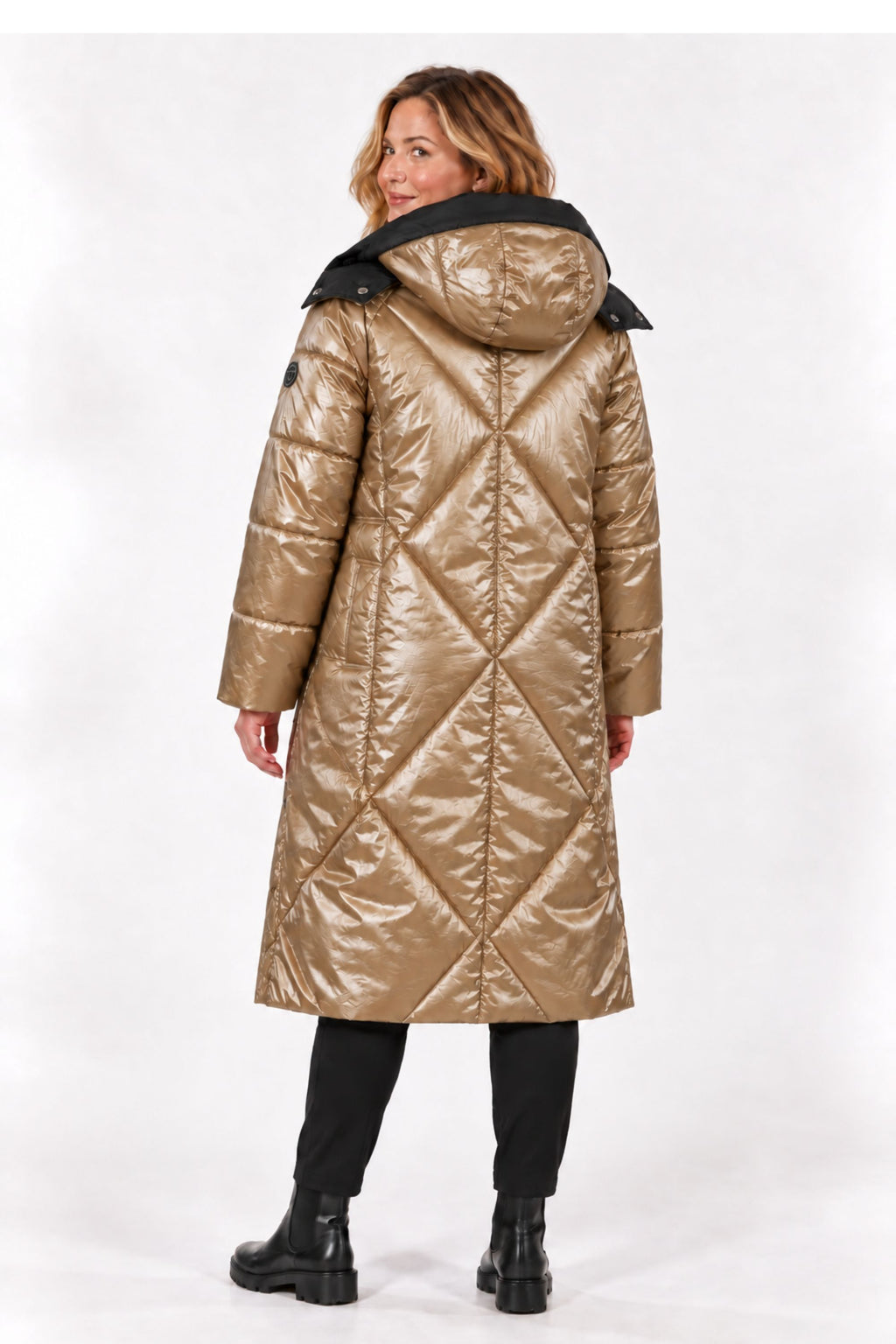 A woman modeling a long, shiny quilted puffer coat in metallic gold with a black hood, paired with black pants and boots, photographed against a plain white studio background.