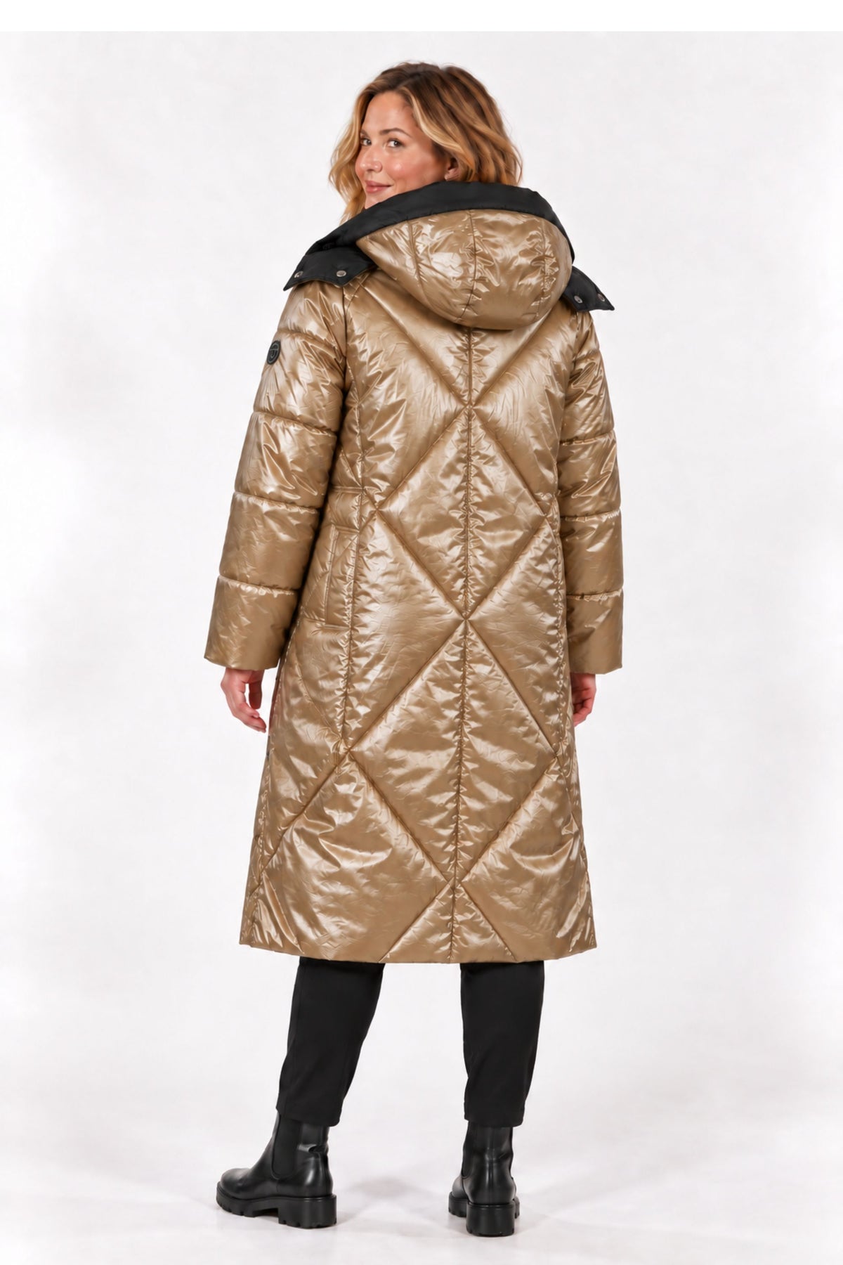 A woman modeling a long, shiny quilted puffer coat in metallic gold with a black hood, paired with black pants and boots, photographed against a plain white studio background.