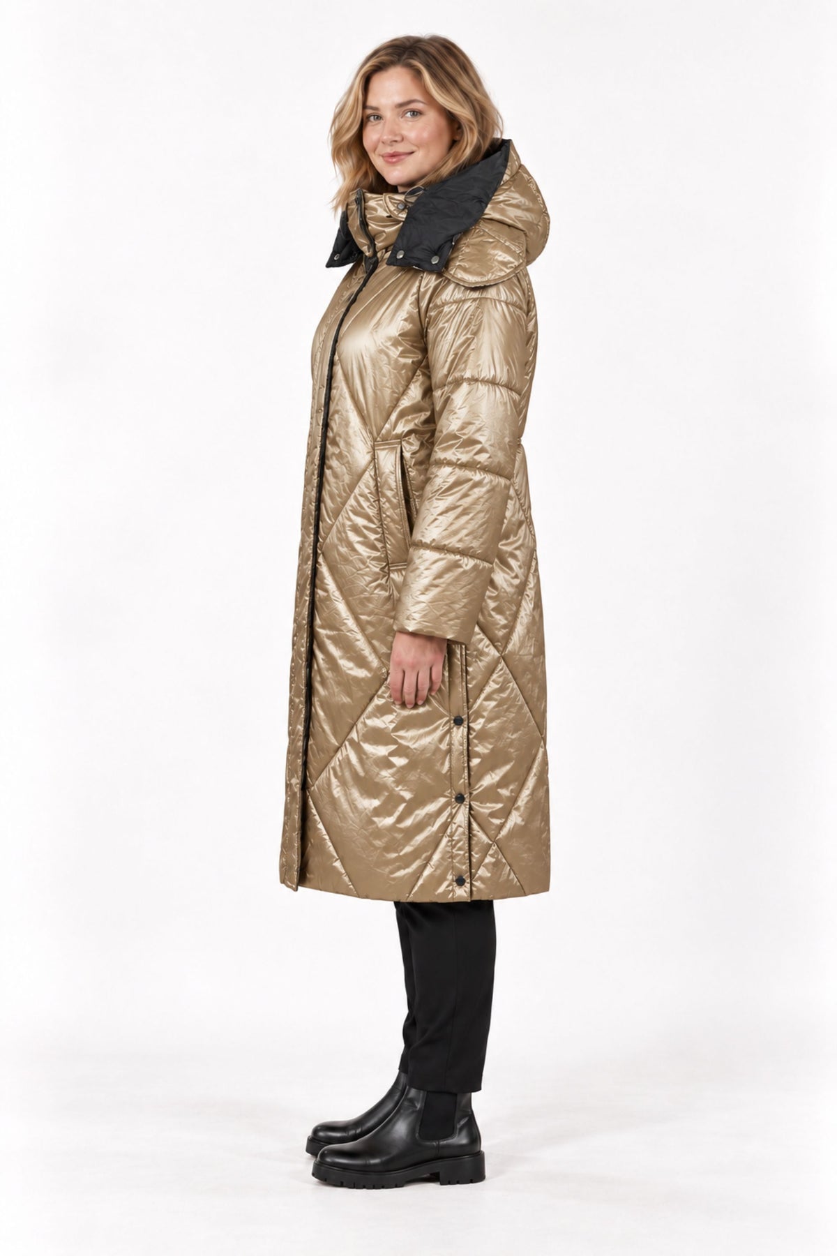 A woman modeling a long, shiny quilted puffer coat in metallic gold with a black hood, paired with black pants and boots, against a plain white studio background.